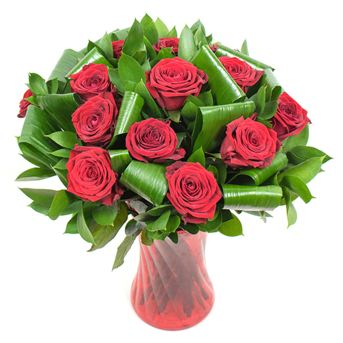 A sumptuous, rounded bouquet of about a dozen richly hued crimson roses commands attention, each bloom perfectly open with tightly spiraled, velvety petals that deepen in tone where light meets shadow. The roses are nestled into an abundance of dark green foliage: compact, flat leaves provide a dense, leafy foundation while broader glossy leaves have been artfully rolled and folded into dramatic spirals and cylinders that arch and loop like green ribbons. These sculpted leaves bring a contemporary, almost architectural accent to the otherwise classic rose silhouette, adding height, depth, and a gentle tension between soft curves and crisp edges. The stems are arranged in a tall, translucent ruby-red glass vase whose subtle ribbing or twisted pattern creates vertical highlights and mirrors the bouquet's passionate red hues; the vase's narrow waist and flared mouth offer a poised counterpoint to the lush floral dome. Photographed against a clear white background, the colors read with brilliant contrast: the deep crimson feels intimate and intense, the greens rich and grounding, and the glass gleams with reflected light. One can almost sense the familiar, powdery scent of fresh roses - an olfactory memory suited to anniversaries, Valentine's Day, or a treasured romantic gesture - and feel the careful hand of a florist shaping each leaf and bloom. In the context of North Watford, I picture this arrangement brightening a table near Cassiobury Park on a cool morning or arriving as a considered surprise to a terrace home off St Albans Road, its combination of tradition and modern leafwork making it equally appropriate for classic and contemporary interiors.