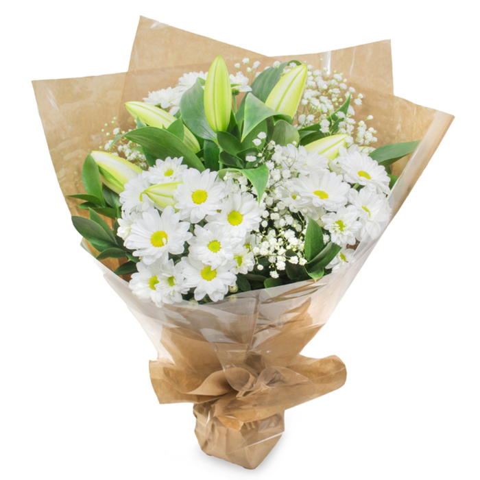 This hand-tied bouquet of white and green flowers is shown upright against a clean white background, every stem visible as though it has just been finished in a North Watford flower shop. The main focus is on generous heads of white daisy-like chrysanthemums, their petals forming tidy layers around bright yellow-green centres that draw the eye inward with a soft, luminous effect. These daisies are nestled closely together, creating a rounded, cushioned look that feels full yet gentle, ideal for placing on a side table or by a framed photograph in a quiet room. Interspersed between them are several tall, elegant lily buds, their forms slender and sculptural, with outer petals shaded in pale green and tips that fade to creamy white, suggesting fragrant blooms that will gradually open in the days after delivery. Threaded delicately through the arrangement are fine sprays of white gypsophila, or baby's breath, the tiny blossoms clustering like a light mist that softens the stronger shapes and adds a sense of airiness. Around the flowers, deep green, glossy leaves provide a rich backdrop, their smooth surfaces catching the light and giving structure, as though cradling the white blooms in a protective embrace. The stems are gathered together and wrapped first in rustic brown kraft paper, whose slightly rumpled texture brings a natural, earthy warmth that feels at home in the residential streets off St Albans Road. This is then enclosed in a layer of clear cellophane, adding a polished sheen while keeping the bouquet secure and fresh. Tied neatly at the base, the arrangement exudes purity, serenity, and understated elegance, making it a fitting choice for sending sympathy across North Watford, whether to a family home, a chapel of rest, or a quiet gathering after the service.