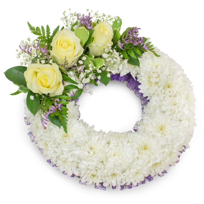 This image shows a measured, compassionate circular wreath designed for sympathy or funeral use, photographed on a stark white surface that highlights every subtle detail. The dominant material is densely grouped white spray chrysanthemums: dozens of small, ruffled heads packed to create a smooth, plush ring whose tactile quality feels soft and reverent. A narrow band of dark lavender-purple mesh or ribbon peeks through at both the inner and outer edges, lending depth and a muted flourish that frames the composition. Placed slightly above the left-hand midpoint is a considered accent cluster where three creamy yellow roses open in gentle bloom; their warm, velvety petals form the arrangement's emotional center. Surrounding the roses are delicate clouds of white gypsophila and tiny sprays of pale purple statice or limonium, which introduce texture and a cool, complementary hue. Greenery - ranging from broad, smooth leaves to fine, fern-like fronds - supports and contrasts the lighter flowers, providing structure and a fresh, living backdrop. The wreath holds a clear, open center symbolising continuity and remembrance; soft, even lighting throws a faint shadow beneath the form, giving it presence against the white plane. As a local florist who has prepared tributes for families near Watford Junction and the High Street, I recognise this as a classic, understated design: calm, consoling, and carefully constructed to express honour and lasting remembrance. Flower Delivery North Watford would describe it as an eloquent, modest choice for a memorial service.