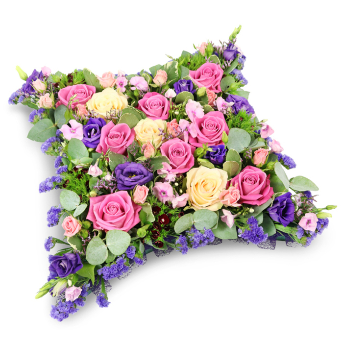 In this detailed funeral tribute, a cushion-shaped arrangement of flowers is shown from above, lying on a pristine white surface that accentuates its colour and texture. The floral pillow forms a soft rectangle with subtly curved corners, conjuring the idea of a resting place fashioned from petals rather than fabric. Across the entire surface, roses in shades of pink and cream mingle with deep purple lisianthus and small filler blooms in lavender tones, creating a harmonious, comforting palette. Fuchsia roses, fully open and luminous, stand out as focal points, their generous heads layered with velvety petals. Nearby, softer blush and pastel pink roses weave through the design, while creamy pale yellow roses introduce a gentle warmth reminiscent of quiet sunrise light over North Watford's peaceful streets. Among them, rich purple lisianthus blooms provide contrast, their slightly ruffled petals bringing a velvety, almost satin-like texture to the design. Finer elements, such as delicate spray roses and tiny clustered blossoms of statice or limonium, add an intricate lacework of shape and colour, ensuring the cushion looks completely full and lush from every angle. Framing and supporting the flowers is a generous collar of foliage: rounded, silvery-green eucalyptus leaves lend a muted, elegant tone, while darker, fine-textured greenery with a fern-like character deepens the overall structure. The arrangement appears carefully balanced, with colour and height evenly spread, suggesting the thoughtful craftsmanship of a specialist in sympathy tributes serving bereaved families in North Watford. It feels like a quiet, tender way to honour a loved one's memory during a funeral, memorial, or graveside farewell.