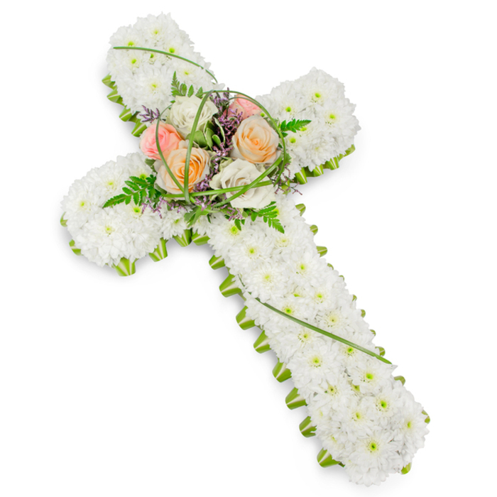 A traditional Christian floral cross, crafted with meticulous care, appears against a seamless white background, inviting quiet contemplation. The body of the cross is entirely swathed in tiny white cushion chrysanthemums, their rounded petals pressed close together to form a gently textured, almost quilted surface. This field of white stretches across the horizontal arms and down the long vertical stem, evoking a sense of peace, innocence, and serenity often sought in funeral flowers for services across North Watford. To frame this softness, a crisp outline of fresh green foliage and neatly arranged floral edging runs around the perimeter of the cross. This vivid green contour hugs each curve and angle, creating a clear, well-defined silhouette that stands out against the neutral backdrop while still feeling natural and soothing. At the central point where the arms intersect, a tasteful spray of larger blooms adds a warm focal point. Several roses in shades of soft peach, pale pink, and creamy ivory nestle together, their velvety petals unfolding in layered rosettes. They are surrounded by lacy fern fronds, slender blades of looping grass, and small, airy accents of purple filler flowers that slip between the petals, adding gentle depth and dimension. The lighting is bright yet soft, with no dramatic shadows, giving the image a calm, contemplative mood. This type of cross would be a meaningful choice for a family in North Watford wishing to honour a loved one's faith and memory, whether placed beside a coffin during a church service or laid tenderly at a graveside as a final farewell.