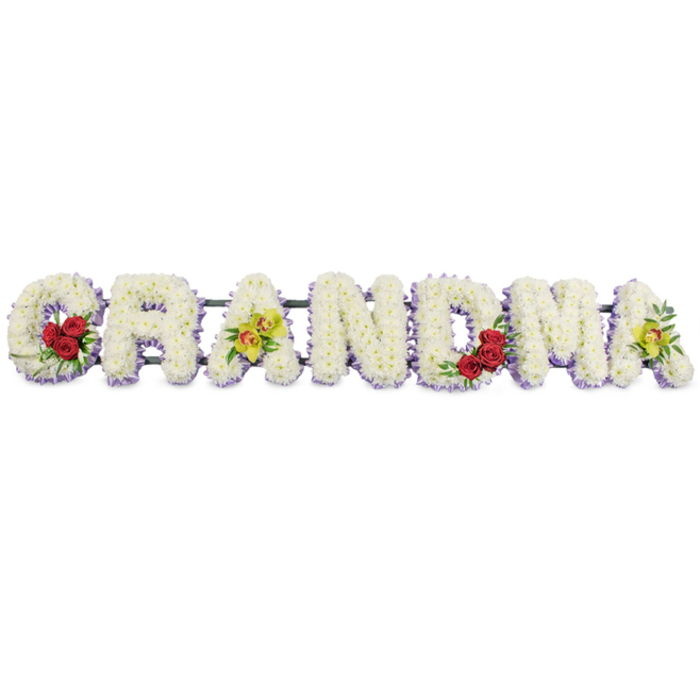 In this photograph, a large sympathy tribute spells out GRANDMA in elaborate floral letters, arranged in a straight line as if ready to be placed beside a coffin or on display at a memorial gathering in North Watford. The backdrop is pure white, eliminating distractions and placing full focus on the skill of the floristry. Every letter is formed from a thick covering of small white chrysanthemums or similar button blooms, so densely grouped that the individual heads merge into a continuous, cushion-like texture. The impression is soft and comforting, inviting the mind to imagine the cool, fresh feel and gentle scent of newly opened flowers. Along the lower edge of each letter, a delicate edging of pale lavender or light purple flowers peeks out, as though the white petals are resting on a soft lilac shadow. This subtle border gives definition and a hint of colour, reminiscent of early evening light over the nearby parks and streets of North Watford. Bolder accents punctuate the tribute in just a few key places. On the lower curve of the opening G, three rich red roses open in a tight cluster, surrounded by dark green leaves that add depth and contrast. The first A displays two yellow-green cymbidium orchids along its bar, their exotic faces with reddish-brown markings suggesting individuality and character. The centre letters R, N, and D remain purely white with their lavender halo, calm and solemn, before the M introduces another trio of red roses on its lower right, echoing the love expressed at the start. The final A mirrors the first, again with two vivid green orchids, visually tying beginning and end. All letters are aligned on a narrow, dark metal frame that discreetly supports the weight of the blooms. The design reads as a heartfelt farewell to a much-loved grandmother, a floral tribute that could accompany the family from the funeral procession through to a quiet moment of reflection after the service.