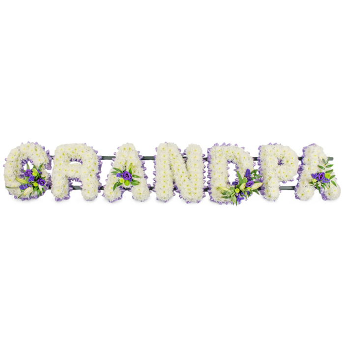 A touching sympathy tribute in the photograph spells out the word GRANDPA in large capital letters, created as a continuous row of floral letters on a concealed frame, ready for display at a funeral or cremation service in North Watford. Each letter is meticulously covered in compact clusters of white chrysanthemums or carnations, giving the surface a thick, cushion-like feel. The overlapping petals create delicate shadows and a gentle shimmer of white-on-white tone, echoing purity, remembrance, and a sense of calm. Across this pristine base, the florist has woven in small groupings of deep purple and soft lavender blooms, supported by glossy green foliage. These coloured accents appear purposefully within key areas: tucked into the lower curve of the G, nestled in the centre of the first A, adding depth within the upright line of the D, glowing softly within the bowl of the P, and enhancing the final A. The purple tones bring just enough contrast to express affection and quiet strength without disturbing the overall serenity. A narrow band of violet ribbon edges parts of the letters, giving the arrangement a neat profile and gently echoing the purple flowers. All of the letters are mounted on a slim, dark green support bar, mostly hidden behind the blooms, ensuring the tribute stays straight when placed at a graveside near North Watford Cemetery or in a local church along St Albans Road. The design feels deeply personal, as if created by a seasoned North Watford florist for a family wanting to say a heartfelt, dignified goodbye to their grandfather in a single, meaningful word.