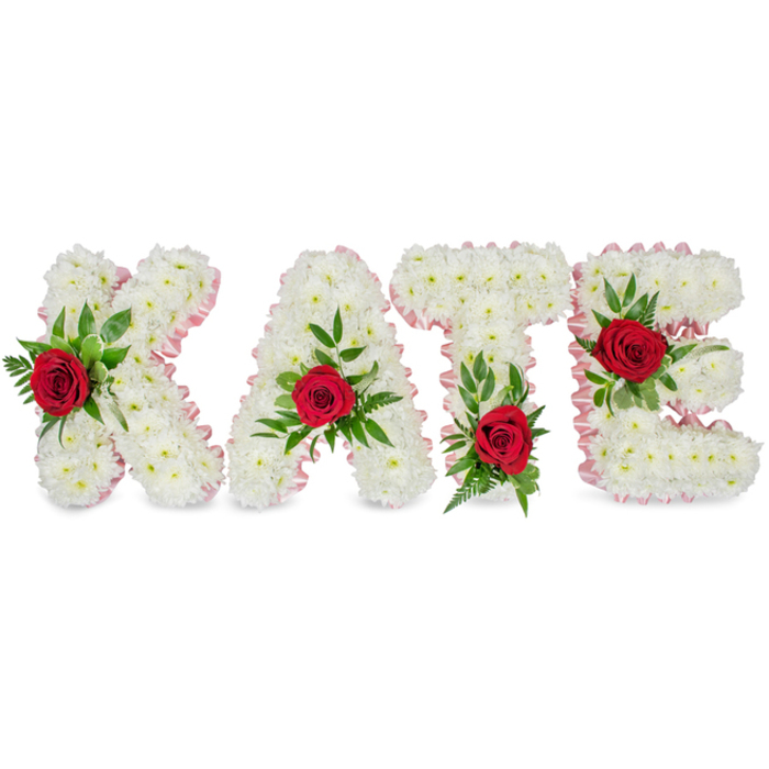 A solemn, beautifully detailed floral name tribute displaying KATE in large, solid block letters against a stark white backdrop, designed with the precision of a seasoned florist. Each of the four letters is densely covered with small, immaculate white chrysanthemums whose rounded faces create a sculpted, voluminous texture reminiscent of soft down or summer clouds. The perimeter of every letter is trimmed with a subtle pale-pink satin ribbon, pleated gently to form an understated frame that adds a whisper of warmth to the otherwise pristine composition. Anchoring the centre of each letter is a single rich red rose: positioned on the lower left vertical stroke of the K, atop the triangular summit of the A, at the base of the left arm of the T's crossbar, and on the middle horizontal bar of the E. These crimson roses introduce a deep, velvety contrast and are tucked into sprays of fresh green foliage - broad glossy leaves and slender fern fronds - which provide structural support and a verdant backdrop to the blooms. The arrangement conveys both dignity and intimacy; the white chrysanthemums read as purity and quiet reflection while the red roses convey heartfelt affection. This sort of bespoke memorial lettering is often chosen for funeral services, church remembrances or celebration of life ceremonies in the North Watford area, offering a personal and graceful tribute that photographs and reads clearly from a distance while remaining richly textured up close.