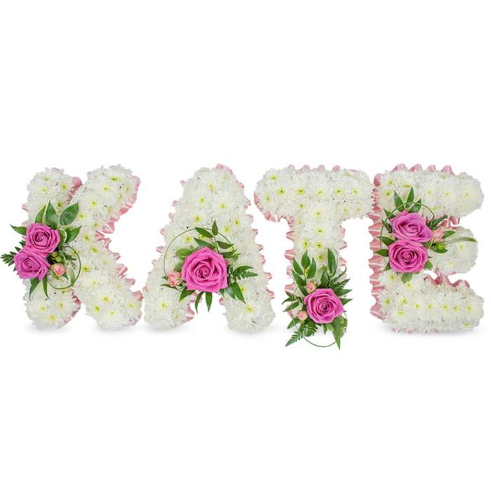 This carefully composed floral tribute spells KATE in substantial, freestanding letters, each letter formed from myriad small, immaculate white chrysanthemums to produce a plush, cloud-like mass of blooms. A whisper of pale-pink satin ribbon follows the internal contours of each character, providing a refined, feminine outline that separates and uplifts the white mass beneath. The arrangement is punctuated by vivid hot-pink roses placed with intention - two on the K's left arm, a single focal rose in the lower sweep of the A, one vivid bloom at the base of the T, and a duet of roses forming a bright accent on the right side of the E - their texture and implied scent bringing warmth and humanity to the composition. Rich greenery - ferny fronds, variegated magnolia-style leaves, sprays of ruscus and delicate eucalyptus - frames the roses and emerges subtly from the chrysanthemum field to add colour contrast and a garden-fresh note. Small pink filler flowers and slender green tendrils soften hard edges and introduce gentle movement across the surface. Presented against a stark white background, the letters appear sculptural and luminous, shadows beneath giving them weight and presence. The overall effect is dignified and tender, an expertly executed memorial or commemorative piece intended to convey remembrance, grace, and heartfelt respect at a funeral or celebration of life.