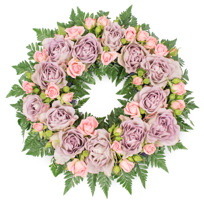 A serene funeral wreath rests flat against a clean white backdrop, its circular form perfectly balanced and full, as though just arranged by a careful hand in a North Watford flower studio. The entire ring is composed of roses in complementary soft tones, with the eye first drawn to the large, fully open blooms shaded in dusty lavender, antique mauve, and cool greyish-purple. These roses display intricate swirls of petals gathered toward deep, gently shadowed centres, giving the design a sense of depth and emotional weight. Interspersed among them are daintier clusters of blush-pink spray roses, many still in bud or only partially open, their smaller, tighter petals offering a youthful contrast to the mature, fully unfurled roses around them. This interplay of bloom stages suggests both remembrance of the past and hope for peace ahead. Beneath and around the flowers lies a thick layer of dark green fern fronds-likely leatherleaf or sword fern-whose finely toothed leaves radiate outward, creating a soft yet structured frame for the wreath. The greenery peeks through between petals, adding texture and a natural rhythm, like the gentle pattern of trees along North Watford's quieter streets. Hints of deeper-toned filler greens and tiny dark buds hide among the roses, subtly increasing the dimensional effect and giving the piece a carefully curated, garden-inspired feel. Soft, even lighting falls across the surface, highlighting the velvety petals and glossed foliage without harsh contrast, so the mood remains calm and contemplative. As a sympathy tribute, this wreath conveys messages of love, honour, and quiet respect, making it a fitting floral gesture for funerals, memorial gatherings, or graveside farewells across North Watford.