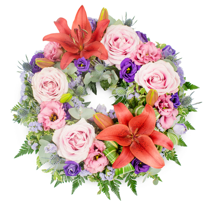 This image shows a lush, perfectly round funeral wreath laid flat against a seamless white backdrop, allowing every colour and petal to be clearly seen. The arrangement is rich and layered, with no gaps, giving a feeling of abundance and care. Two prominent Asiatic lilies in a warm coral-orange hue anchor the design, one towards the upper curve and another near the lower right, their wide, star-shaped blooms displaying long stamens dusted with pollen. Between and around them, clusters of soft pink and lavender-toned roses open fully, their spiralling centres drawing the eye inward and softening the stronger colours. Interspersed among the roses are lisianthus (eustoma) blossoms, some in pale, almost blush pink, others in regal, velvety purple, their delicately ruffled petals adding a gentle, romantic texture. Tiny sprays of lavender-blue filler flowers, similar to statice, thread lightly through the wreath, softening transitions and creating a misty effect. The base is built on a foundation of mixed greenery: rounded, silvery eucalyptus leaves, slender fern fronds, and darker foliage that adds depth. Striking blue-grey eryngium thistles punctuate the design with their spiky, cone-shaped heads, lending a subtle contemporary edge. Near the lower lily, bright green leaves are folded and sculpted, highlighting skilled craftsmanship. Several unopened lily buds peek from the foliage, suggesting the continuation of life beyond loss. The whole wreath feels serene and respectful, the kind of tribute that would be chosen for a funeral or memorial gathering in North Watford, perhaps delivered to a local church or crematorium to honour a loved one with grace.