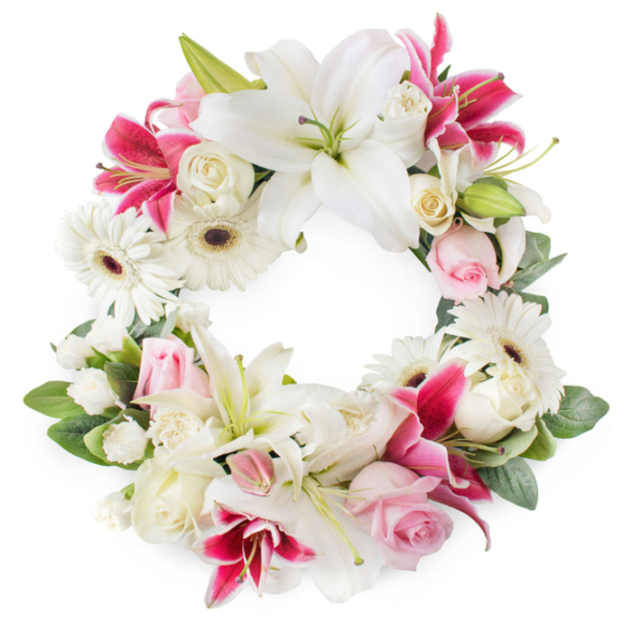 A beautifully balanced circular sympathy wreath is photographed against a pristine white background, its open centre and carefully arranged flowers creating an immediate sense of quiet reflection. The colour story is one of gentle contrast: cool whites and soft blush pinks interlaced with deeper magenta tones, forming a ring that feels at once tender and dignified, perfect for a North Watford funeral or celebration of life. Dominating the composition are large white lilies, their petals wide and slightly recurved at the tips, revealing delicate green centres and bright golden stamens that lend subtle warmth to the arrangement. Threaded between them are rich pink and fuchsia lilies, some with faint speckling on their petals, which bring depth and a more intense note of remembrance without disturbing the overall calm. White gerbera daisies punctuate the wreath with their round, open faces and dark, almost chocolate-coloured centres, bringing a fresh, contemporary touch. Around them, clusters of pale pink roses and creamy white roses are nestled closely together; some blooms are tight and youthful, while others are fully opened, with overlapping petals that suggest long, shared memories and enduring love. Fine-textured white filler flowers-likely spray carnations or similar multi-headed stems-soften the transitions between larger blooms, ensuring the circle remains plush, full, and visually continuous. Everything is anchored by a bed of lush green foliage, with layered leaves providing depth, shadow, and a natural frame to the floral display. This circular wreath, carefully composed by an experienced local florist and suitable for delivery to North Watford funeral homes, churches, and memorial venues, speaks of peace, purity, and the unbroken cycle of love and remembrance.