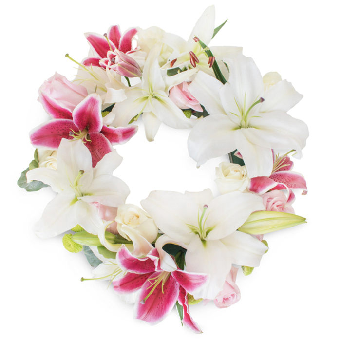 Centered on a pristine white background, a magnificent floral wreath radiates calm and reverence, reflecting the artistry of North Watford's trusted florists. The circular design is lush with brilliant white Oriental lilies, some fully open and arching gracefully, others gently curled as if just roused by morning air. Each white bloom is offset by vibrant Stargazer lilies-a vivid fuchsia, speckled with threads of magenta and edged in soft white-providing exhilarating bursts of colour and infusing the display with the spirited energy found in the heart of North Watford's neighborhoods. Between the lilies, clusters of pale blush and creamy white roses nestle close, their delicately swirled petals lending romance and a soft sense of remembrance, echoing the gentle support of community during times of loss. Subtle sprays of muted green foliage, possibly eucalyptus or lily greens, serve as a grounding element, echoing the tranquillity of the parks and gardens throughout North Watford. The composition's dense, unbroken circle evokes unity and eternal love, making the wreath an ideal centrepiece for memorial services, funerals, or quiet celebrations of a life well-lived. Every detail-down to the harmonious mix of colour, texture, and form-reflects the compassion and expertise that define local craftsmanship, offering profound comfort to families and loved ones from North Watford and beyond.