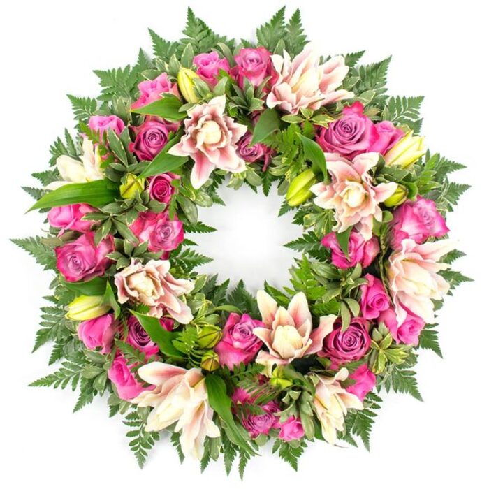 Set against a plain white studio background, this circular funeral wreath appears full, composed, and richly layered, as though freshly created for a service in North Watford. The eye is first drawn to the sweeping band of roses that circle the frame: velvety blooms in saturated fuchsia and magenta mingle with softer pinks, creating a rhythm of colour that moves seamlessly around the ring. Some roses are open wide, revealing their intricate spirals of petals, while others remain semi-closed, lending a sense of tenderness and newness. Interwoven throughout are generous double lilies, their petals a blend of creamy white, blush pink, and whisper-soft peach, with a few petals tipped or veined in deeper pink. Several lily buds, in vibrant yellow-green, rise slightly above the blossoms, adding vertical interest and a feeling of life continuing. The entire floral circle is underpinned by a lush collar of greenery: dark emerald fern fronds splay outwards like a soft fringe, while glossy leaves fill the inner spaces, providing contrast and structure. The composition is deliberately symmetrical yet never stiff; it feels like a carefully considered gesture of love and remembrance. The lighting is even and gentle, so each petal texture and each curve of foliage is visible without harsh shadow. This kind of wreath, with its romantic pink palette and tranquil character, would be well suited to a celebration of life at a local church or gathering place, expressing sympathy with grace and quiet beauty.