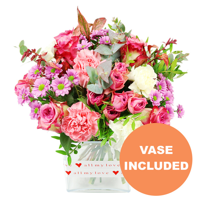 In this product-style image, a vibrant mixed bouquet stands out strikingly against a clean white backdrop, every bloom clearly visible in a clear, rectangular glass vase. The arrangement feels full and generous, dominated by roses and carnations in a harmonious spectrum of pinks. Large, fully opened roses in rich fuchsia and deep blush command attention near the centre, their layered petals curling softly at the edges. Some of the roses display darker, almost magenta tips, while others are a more uniform pastel pink, evoking a sense of tenderness and romance. Nestled amongst them are ruffled carnations in coral, salmon, and peach-pink tones, their frilly petals adding texture and a slightly playful, ruffled look. Interspersed throughout are clusters of smaller daisy-like chrysanthemums in soft lavender and purple shades, each with a bright lime-green centre that provides a lively contrast and draws the eye deeper into the arrangement. Creamy white flowers are carefully placed to prevent the design from becoming too saturated, giving moments of calm between the bolder colours. The greenery is abundant and thoughtfully layered: rounded eucalyptus leaves in silvery green share space with darker glossy foliage and a scattering of reddish-brown leaves, which lend a subtle, seasonal or romantic depth. The flowers are presented in a simple yet stylish clear glass vase with rounded corners, wrapped at the base in a white decorative band. This band is printed with repeating red hearts and the phrase all my love in flowing red script, reinforcing the sentiment behind the bouquet. In the bottom right corner, an orange circular overlay with crisp white lettering proclaims VASE INCLUDED, indicating that the arrangement is ready to display as soon as it arrives. The overall effect is of a professionally crafted bouquet you might have delivered in North Watford, ideal for local celebrations, anniversaries, or Mother's Day surprises.