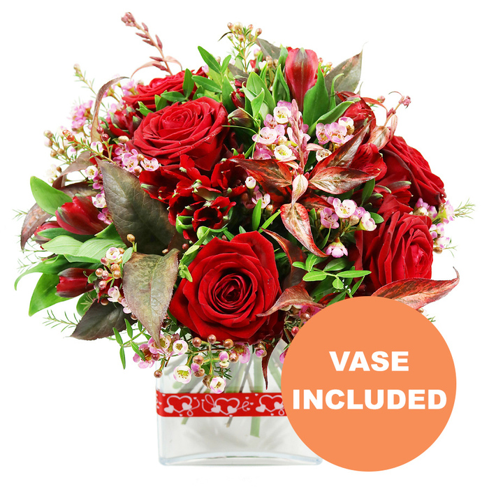 A sophisticated, full-bodied floral arrangement in rich shades of red and green stands out crisply against a pure white background, the sort of stylish bouquet you might send to a loved one in North Watford for an anniversary dinner or Valentine's evening at home. Dominating the composition are several large velvety red roses, each bloom at a slightly different stage, from tight, promising buds to more open flowers revealing layered petals that curl softly at the edges. These signature roses are surrounded by clusters of smaller, deep red flowers similar to alstroemeria, their multiple petals contributing a ruffled, textural counterpoint that fills the spaces between the larger blooms. Woven delicately throughout the bouquet are airy sprays of tiny filler flowers, likely waxflowers or limonium, in pale pinks and whites that bring a gentle, romantic lightness and help to break up the intensity of the darker reds. Encircling the flowers, a generous collar of greenery in varying shades and shapes creates a sense of natural abundance: some leaves are broad and glossy, others slender and more feathery, mimicking the layered textures you might see in well-kept front gardens off St Albans Road. Adding a sophisticated twist, variegated foliage in burgundy and reddish-brown tones is strategically placed, the subtle veining and warm colour echoing the deep crimson roses and enhancing the bouquet's dramatic, almost couture styling. The flowers are arranged in a clear rectangular glass vase with soft edges, clean lines that emphasise the modern feel of the design, while the visible stems give a reassuring sense of freshness. A red ribbon, patterned all around with interlocking white heart outlines, is neatly tied near the base of the vase, gently reinforcing the message of love and affection. In the bottom right corner of the image, a bold orange circular label with white uppercase text states VASE INCLUDED, clearly highlighting that this elegant, gift-ready arrangement is delivered complete with its own vase, perfect for setting straight onto a dining table or windowsill in North Watford.