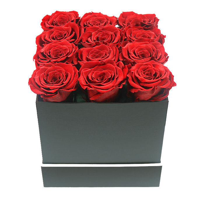 A refined close-up photograph presents a square black gift box overflowing with the saturated beauty of twelve ruby red roses, styled in a way that feels both modern and timeless, much like the quiet residential streets of North Watford. The roses are arranged in a precise 3x4 pattern, each bloom perfectly aligned so that their rounded heads create a level, velvety canopy of crimson. Every rose appears at its peak, the petals unfurling in dense, layered spirals that suggest carefully preserved or 'eternal' roses chosen for their resilience and long-lasting form. Their colour is a deep, luxurious red with delicate highlights along the petal edges where the soft lighting lands, revealing subtle shifts from rich garnet to slightly brighter scarlet. The blooms sit snugly within a matte black, square box that feels almost architectural, with clean, unwavering lines and an unadorned surface. Around the lower third of the box, a slim white band runs in a continuous ring, creating a simple yet striking contrast-like a narrow frame that draws the eye back up to the roses. The background is pure white, making the arrangement appear as if it's floating in space, and preventing any visual clutter from distracting from the floristry. Gentle, even lighting softens the shadows at the base of each rose and along the box edges, adding depth without losing clarity. This sophisticated composition conveys enduring romance and high-end gifting, equally suited to a Valentine's surprise delivered near North Watford's local cafés or a statement anniversary present arriving at an office close to St Albans Road, where its bold elegance can quietly transform a desk or side table.