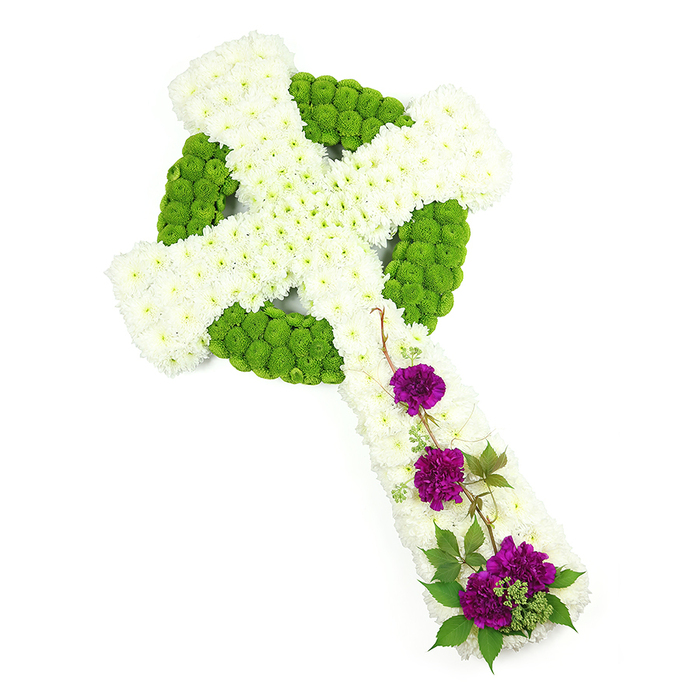A beautifully detailed Celtic cross funeral arrangement is displayed diagonally across a bright white background, allowing every element of the floral artistry to be appreciated. The cross itself has a strong, sculpted silhouette, with the vertical and horizontal arms clearly defined by a dense covering of small, snow-white chrysanthemum heads. These blossoms are arranged so closely that they form a pillowed, almost quilted surface, gently rounded to give the design a three-dimensional presence. Encircling the centre of the cross, and threading through the design in subtle bands, sit compact lime-green button chrysanthemums. Their vivid chartreuse colour draws out the Celtic ring and hints at interlaced knot patterns, echoing ancient carved crosses often associated with Irish and Scottish heritage. The green blooms nestle between the white flowers, adding texture, depth, and a sense of living freshness, like new growth against weathered stone. On the lower length of the cross, three prominent clusters of deep purple carnations add drama and emotional warmth. Their petals are abundant and frilled, layered in rich shades of plum and magenta, and they are framed by trail-like sprigs of dark green foliage, perhaps ivy or similar, that curl and weave gracefully across the white chrysanthemum base. The lighting is high and even, emphasizing the clarity of the colour palette-pure white, vibrant green, and regal purple-and highlighting the meticulous craftsmanship. Created as a thoughtful sympathy tribute for families around North Watford, this floral Celtic cross would sit beautifully beside a coffin, at the front of a chapel, or placed on a grave, quietly expressing faith, peace, and enduring love.