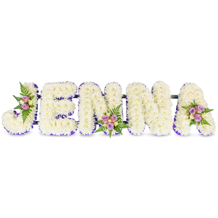 A tender, personalised memorial piece spells the name JENNA in bold capital letters, each letter built from a dense mass of creamy-white chrysanthemum florets or similar small white flowers to create a soft, cushiony texture that feels both gentle and substantial. The inner rims and bases of the letters are edged with a refined band of vivid purple-lilac ribbon or tiny contrasting blooms, which adds elegant definition and a subtle visual contour against the pure white field. Small, artful clusters of blush and lavender spray roses, accompanied by a few lighter cream or pale yellow roses and feathery dark-green fern fronds, are thoughtfully placed on the upper left curve of the 'J', the central base of the second 'N', and the upper right sweep of the 'A', punctuating the name with moments of warmth and living colour. Thin, unobtrusive dark bars join the letters horizontally, creating a secure, cohesive structure suitable for display on an easel at a funeral, wake or chapel. Under soft indoor lighting the whites read as serene and the purple trim provides a restrained dignity; the roses lend a gentle fragrance and the ferns add depth and a natural finish. As a design well suited to services around North Watford - near Cassiobury Park, the town centre and local funeral homes - this floral name tribute expresses respect, remembrance and the quiet craftsmanship of a local florist, offering a deeply personal and beautifully made farewell.