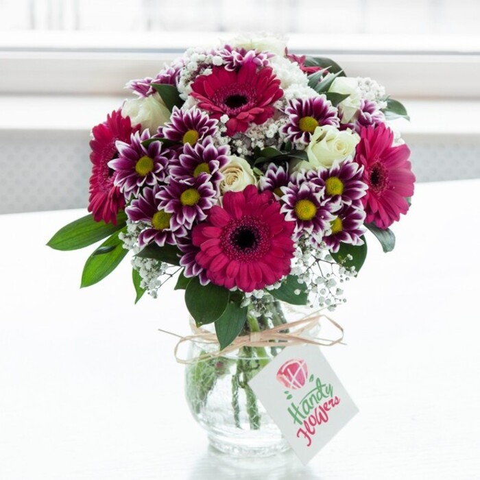 A lush, joyful flower arrangement stands center frame in a squat, clear glass vase, bathed in soft, natural light that gives the impression of a bright North Watford morning. The bouquet bursts with colour and texture: large gerbera daisies in intense fuchsia and magenta radiate from the middle, their broad petals forming perfect circles around dark, almost chocolate-colored centers that add depth and drama. Encircling them, striking purple-and-white chrysanthemums display layers of slender petals; each bloom shows a deep purple hue at the tips that gently fades into clean white nearer the base, drawing the eye inward to cheerful yellow centers that glow like tiny suns. Interwoven among these bolder flowers are creamy white roses, some fully open and cupped like porcelain, others still in bud with their petals wrapped closely around each other, hinting at the promise of days of freshness ahead. Wisps of airy baby's breath, with their countless tiny white blossoms, drift through the arrangement like a fine veil, creating a soft, romantic counterpoint to the strong magenta and purple tones. Rich, dark green foliage frames the flowers, providing structure and a garden-fresh backdrop that makes the colours appear even more vivid. The glass vase, with its subtle texture, reveals the straight, neatly trimmed stems inside and is finished with a rustic touch: a loop of natural twine tied into a simple bow around the neck. From this twine hangs a small, rectangular white gift tag showing a delicate pink rose motif and gentle script, suggesting a thoughtful handwritten note. Behind the bouquet, the background remains bright and softly out of focus, as if it were a white windowsill or table in a tidy home near North Watford's parks and residential streets. The composition feels like a freshly delivered gift, the kind Flower Delivery North Watford might bring to celebrate a birthday, express heartfelt thanks, or simply add a burst of happiness and colour to an otherwise quiet room.