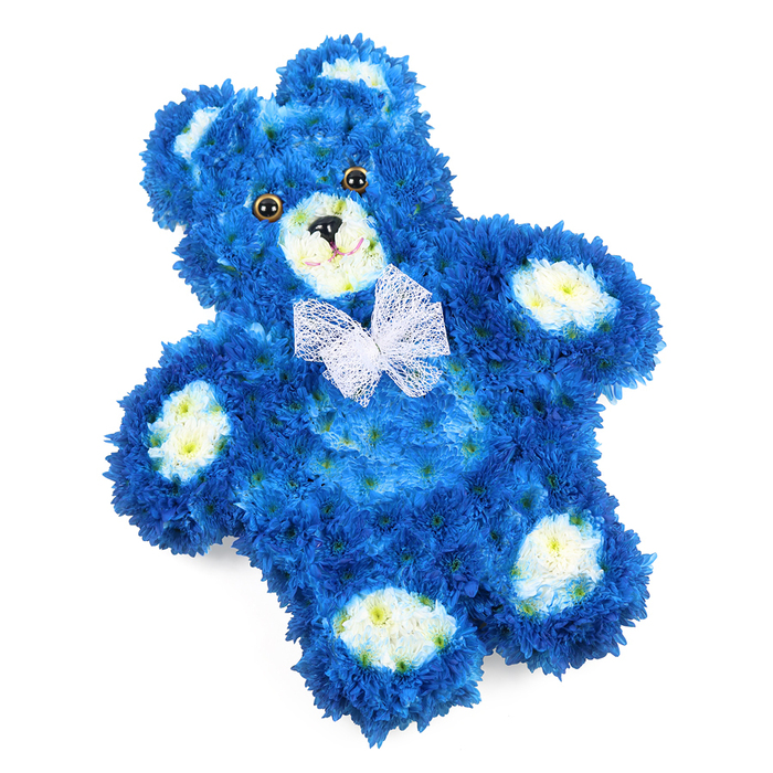 This image shows a lovingly crafted floral teddy bear tribute, resting on a clean white background so that every petal and contour stands out. The bear is predominantly royal blue, its entire form densely covered with small, rounded blossoms, likely chrysanthemums, placed so closely together they resemble a soft, velour coat. Each curve is carefully shaped: a full, rounded head; compact ears set slightly to the sides; a gently protruding tummy; and short, comforting limbs that suggest a toy meant to be held. Light falls evenly over the design, emphasizing the richly saturated blue and the natural shadows that give the bear volume and depth.

In contrast, creamy white and pale yellow flowers mark the bear's key features. The rounded snout is softly highlighted in these paler blooms, giving the face definition and a sense of warmth. Matching light patches nestle inside each ear and form the four oval pads on the hands and feet, echoing the detailing of a classic stuffed teddy. Dark round eyes, slightly glossy, sit above a neat black triangular nose, while a thin black line curves just beneath, hinting at a gentle, reassuring smile that feels especially comforting in times of loss. Around its neck, a sheer white mesh or organza bow is tied in a delicate bow, the loose ends spilling lightly over the chest.

For families in North Watford, this floral teddy bear could be chosen as a touching tribute at a child's funeral in the local churches near Bushey Mill Lane or as a memorial display for a baby boy. The blue tones quietly speak of serenity and remembrance, while the teddy bear shape recalls innocence, bedtime comforts, and love that endures. Its detailed craftsmanship reflects the care of a local florist creating something deeply personal for a moment of farewell.