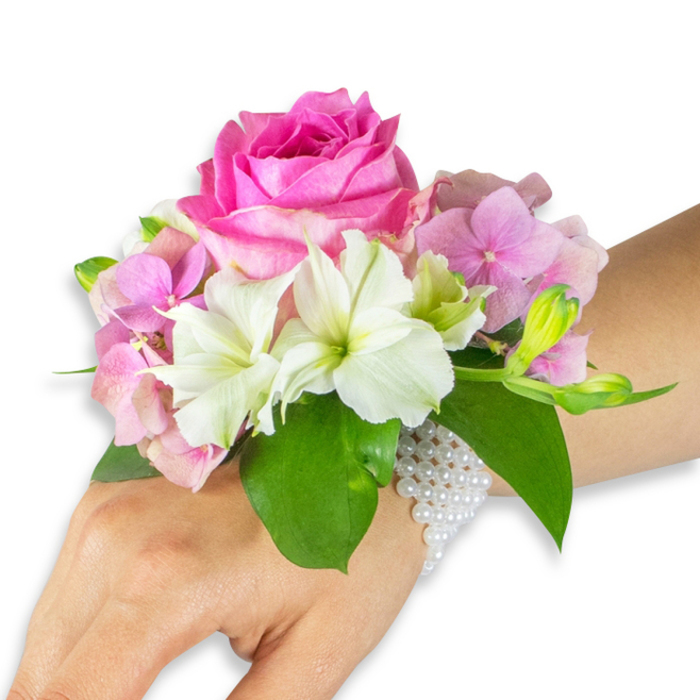 Seen against a pristine white background, this wrist corsage is presented on a left hand and forearm of a light-to-medium skin tone, each element described with the care of an experienced local florist from Flower Delivery North Watford. The focal point is a large, fully bloomed hot-fuchsia rose whose layered petals form a rich, velvety centre that captures soft light and hints at a gentle fragrance. Nestled beneath and around the rose are clusters of pale blush-pink hydrangea florets that add plush texture and a romantic, airy fullness. Interspersed with the hydrangea are crisp white alstroemeria (Peruvian lilies), their four-to-six petaled faces edged with subtle green veins that provide delicate contrast and botanical finesse. Toward the outer edge of the corsage a tidy cluster of pale green alstroemeria buds and a few smaller, barely opened pink buds suggest unfolding beauty, while broad, glossy emerald leaves cradle the arrangement and ground the palette. The entire composition is attached to the wrist by an elegant, multi-strand bracelet of small white pearls, forming a fine, netted band that drapes in a refined curve and extends partially down the back of the hand. The studio lighting is soft and even, emphasizing the tactile softness of the petals and the cool sheen of the pearls. This carefully balanced wearable bouquet would suit a bride's party, prom or graduation and carries a local sensibility picked up from arranging orders for couples near Vicarage Road and the quieter streets off Hempstead Road.