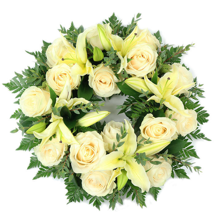 This overhead photograph captures a circular memorial wreath carefully assembled from creamy white roses and pale yellow lilies, set on a plain white background so every texture and tone reads clearly. The arrangement is intentionally dense: approximately a dozen to twenty creamy roses-each with tightly coiled petals and a satiny surface-anchor the piece, their ivory hues ranging from warm cream to soft pearl. Among them, trumpet-shaped lilies in a subtle lemon shade open into graceful curves; some are fully expanded, mapping delicate veins along their petals, while others remain in elongated buds that hint at gradual bloom and add vertical interest. The green base is layered and varied-soft, feathery fern tips create a halo around the flowers and broader, darker sprigs that resemble eucalyptus lend weight and cooler green tones. The contrast between the pale blossoms and the deeper foliage heightens the wreath's visual serenity. The scent one imagines is gentle: the roses offering a honeyed, powdery perfume and the lilies giving a fresher, green note, together creating a soothing presence suitable for times of mourning. Viewed as a gesture of condolence, the circular form symbolizes unbroken remembrance; its quietly refined palette evokes purity, peace, and steadfast love. For residents near Watford Junction or taking a moment in Cassiobury Park, the wreath's composed simplicity will feel familiar and respectfully rooted in the calm character of North Watford.