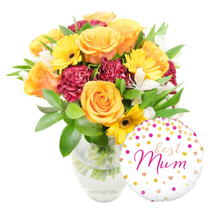 Against a stark white background, a joyful floral composition is displayed in a clear glass vase, the stems and fresh water visible through the transparency and hinting at recent, careful cutting. The bouquet glows with warm tones: large, peach-orange roses dominate with layered, velvety petals that unfold like soft suns; bright yellow gerbera daisies break through with flat, smiling faces and dark centres that draw the eye. Rich burgundy carnations provide deep, tactile texture with their ruffled, compact heads, and delicate white alstroemeria (Peruvian lilies), each petal touched with subtle speckling, thread through the arrangement to soften and brighten. Lush green foliage-broad glossy leaves and finer sprigs-wraps the flowers, delivering both volume and a fresh, leafy scent that evokes the green spaces around North Watford. To the lower right, slightly overlapping the vase and lower blooms, a round white foil balloon adds a celebratory accent: patterned with cheerful gold and hot-pink polka dots and tiny hearts, its centre reads best Mum in an elegant fuchsia script with flowing strokes. The composition reads as a considered gift pairing-flowers with a personalised balloon-perfect for sending from a local shop to addresses near Watford Junction, Hempstead Road, or the surrounding neighbourhoods. It conveys craftsmanship, freshness, and warmth: the tactile contrast of soft rose petals, ruffled carnations, and crisp foliage, finished by a playful balloon that carries the emotional message of appreciation and celebration.