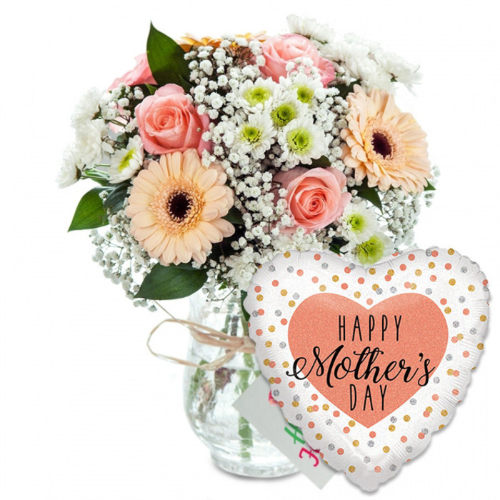 This image shows a thoughtfully coordinated Mother's Day gift set, styled with the gentle elegance you might find in a North Watford florist's window. Everything is arranged on a plain white background, which acts like soft studio lighting, drawing attention to the colours and shapes without distraction. On the left sits a rounded mixed bouquet standing proudly in a clear, jar-style glass vase. The glass reveals the green stems gathered together and gives a glimpse of the water line, reinforcing the sense of freshness and recent preparation. Around the vase neck, a narrow piece of light brown raffia or twine is tied in a loose, simple bow, adding a touch of rustic, handcrafted charm familiar to those who love local, independent shops near the North Watford station area. The bouquet itself is a lush mix of spring blooms. Several peach and light pink roses open gently, their layered petals curling outwards in a classic spiral. They are joined by two striking peach gerbera daisies, each with a bold, dark brown centre that anchors the softer surrounding tones. Nestled between these focal flowers, delicate clouds of white baby's breath introduce an airy, almost frothy texture, while smaller white chrysanthemum heads, each with a subtle pale green centre, create fullness and a sense of movement. Rich green foliage frames the bouquet, slipping in and out between blossoms, making the arrangement feel both polished and natural, like something freshly brought in from a Watford garden. To the lower right, overlapping the base of the vase, a glossy heart-shaped foil balloon becomes a second focal point. Its clean white surface is dotted with sparkling gold and silver circles, reminiscent of confetti scattered at a celebration. At its centre sits a soft peach or light coral heart filled with the message HAPPY Mother's DAY in crisp black lettering, combining a bold, modern font with flowing handwritten script. Just behind the balloon, a corner of a small white greeting card peeks through, underlining the theme of thoughtful gifting and making the whole scene feel like a complete Mother's Day surprise, ideal for doorstep delivery across North Watford.