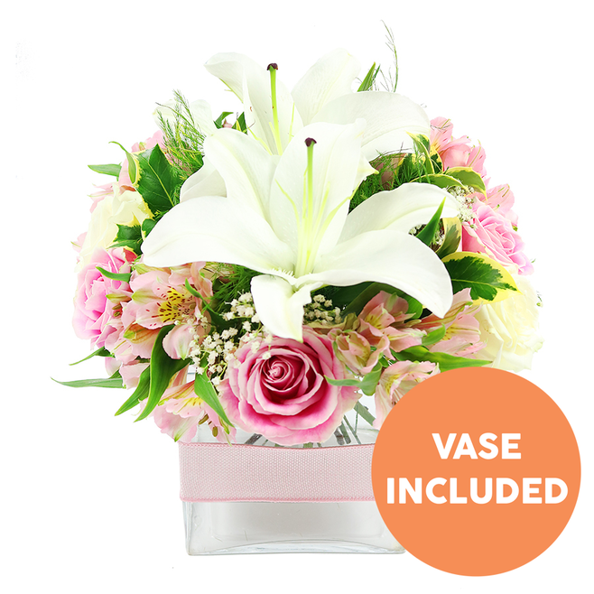 This image shows a beautifully balanced floral centerpiece placed against a crisp white background, the clean setting allowing every petal and leaf to stand out. The arrangement is built around two impressive white Oriental lilies that rise from the heart of the design, their large, curved petals fully open and glowing softly, each bloom showcasing distinct brown-tipped stamens and a central pistil that adds a touch of dramatic contrast. Around these lilies, clusters of pink roses are artfully grouped, some emerging as tight spirals of promise, others already opened into layered, romantic rosettes that exude warmth and tenderness. Between the roses and lilies, alstroemeria blooms in shades of blush and white lend a delicate, painterly pattern, their slender petals lightly streaked and speckled with yellow, creating subtle bursts of colour. Wisps of white baby's breath appear throughout, like small clouds of froth, filling gaps and softening edges. A rich variety of greenery supports the entire composition: broad, glossy leaves catch the light, while finer, feathery foliage tucks in and around the blooms, adding volume and a natural, garden-inspired feel reminiscent of the leafy paths near North Watford's local parks. The flowers are gathered firmly into a low, square clear glass vase, which reveals neatly trimmed stems and clear water, suggesting recent, careful preparation typical of a professional florist. A pale pink ribbon is wrapped horizontally around the vase, tied snugly to introduce a subtle decorative accent without overpowering the flowers. In the bottom right corner of the image, a vivid orange circular label declares VASE INCLUDED in bold white capital letters, clearly indicating that the container forms part of the arrangement. Overall, the bouquet conveys refinement, freshness, and an inviting sense of celebration, ideal for gracing a North Watford dining table or welcoming guests into a bright hallway.