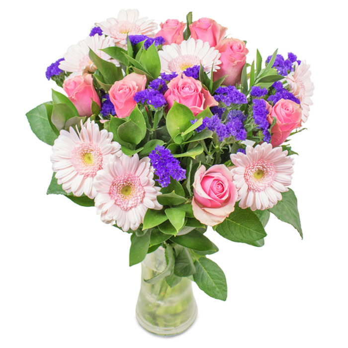 An elegant floral arrangement of soft-to-medium pink roses and pale pink Gerbera daisies, set among clusters of bright purple statice and rich green foliage, all gathered in a clear glass vase with visible stems and water. The roses show varied stages of opening-some tightly furled, others just beginning to reveal their classic spiral centers-creating a sense of living movement and anticipation. The Gerbera daisies contribute broad, layered petals and slightly lighter centers that catch the light and lend a luminous softness to the bouquet. Sprays of purple statice or limonium weave through the composition, their papery texture and saturated violet tone providing a spirited contrast that prevents the pastel palette from feeling one-dimensional. Deep, glossy greenery forms a generous underpinning, the leaves giving a fresh, cool scent and a verdant richness that enhances the blooms' colours. The clear vase keeps the presentation honest and relaxed; stems criss-cross in the water, hinting at careful hand-tying beneath the rim. The overall mood balances joyful celebration with soothing restraint-perfect for birthdays, Mother's Day, an anniversary, or simply to bring a touch of spring into a North Watford sitting room near Watford High Street. Arranged with an experienced florist's eye, this bouquet reflects seasonal freshness and a considered composition that invites both sight and scent.