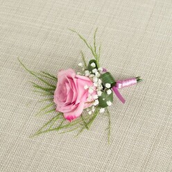 A delicately crafted blush pink rose boutonniere rests on a pale beige, linen-like fabric, its subtle weave visible beneath the flowers like the table coverings often seen at intimate celebrations in North Watford's cosy restaurants and community venues. At the heart of the design, a semi-open rose unfurls in gentle spirals, the inner petals a soft, powdery pink that deepens gradually toward the outer edge, where a slightly more vibrant blush catches the light and hints at a romantic fragrance. Around its base, airy stems of white baby's breath form a soft mist of tiny blooms, each petal catching the light and offering a gentle contrast to the richer rose tones. Wispy, grass-like foliage in a fresh, bright green arcs out behind the central flower, adding movement and a sense of natural ease, while a broader, darker green leaf is folded with precision to wrap around the lower portion, giving the piece a crisp, tailored finish that would look at home on a groom's lapel heading to a ceremony near St Albans Road. The stems are carefully wrapped in a two-toned satin ribbon, blending lavender-pink with a deeper magenta purple, creating a smooth, lustrous band that secures the arrangement and adds a refined touch of colour harmony. Soft, diffused lighting washes over the boutonniere, highlighting the velvety texture of each petal and the gentle shine of the ribbon, while shadowed contours lend depth and realism. This photograph captures the quiet elegance of a classic wedding or prom boutonniere, the kind of handcrafted detail a North Watford florist might prepare for same-day delivery to complement tailored suits and romantic, modern celebrations.