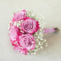 In the heart of North Watford, this expertly crafted bridal bouquet captures the artistry and grace of professional floristry, uniting lush pink and lavender roses in a harmonious dance of colour and form. Each rose reveals a stunning two-tone palette: lavender petals softly glowing at their core and gradually giving way to striking magenta edges, creating a captivating visual depth. Lace-like baby's breath, pure and white, weaves between the larger blooms, bringing both airiness and a gentle spring fragrance that subtly evokes evenings along Cassiobury Park. The bouquet's handle, swathed in a rich plum satin ribbon and encircled by luminous white pearls wound with painstaking care, both dazzles in its detail and invites a loving touch on a momentous occasion. Beneath the decorated wrapping, fresh green stems hint at the bouquet's authentic roots. Placed gently atop a textured, pale linen surface, the image feels softened by natural light, as though caught just before the vows at a sunlit Watford venue. This arrangement, conceived with local flair and a nod toward enduring romance, is ideal for North Watford weddings or milestone celebrations, blending modern elegance with classic sentiment in every petal and pearl.