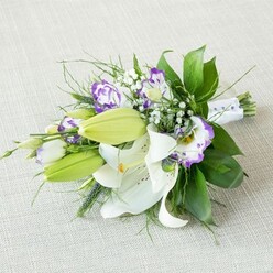 Resting at a soft diagonal across a light beige, subtly textured fabric, this elegant bouquet looks as though it has just been placed down by a bridesmaid pausing for breath before the ceremony. The arrangement centres on several grand white Oriental lilies, their star-shaped blooms fully open so that every velvety petal, gentle speckle, and deep yellow-orange stamen is clearly visible. Alongside them, smooth, pale green lily buds reach forward like quiet promises, still closed but already hinting at the fragrance and drama to come. Twining through the lilies are Lisianthus blossoms, each bloom a small study in contrast: creamy white petals ruffled at the edges and tipped in tones of lilac, lavender, and vivid purple, with some flowers showing deeper colour concentrated at their centres. Sprigs of white baby's breath break up the stronger blocks of colour, sitting like tiny stars in the spaces between, giving the bouquet a romantic, cloud-like softness. The greenery is deliberately varied, with strong, glossy dark green leaves providing structure at the base, then lighter, more delicate foliage and a few airy, grass-like strands reaching outward to create a sense of effortless movement. All the stems are drawn neatly together and wrapped with a smooth white ribbon, finished with discreet pearl-like beads that catch the light just enough to signal something special, perhaps a wedding in North Watford or an anniversary dinner near St Albans Road. The overall atmosphere is one of freshness and quiet celebration, the kind of carefully crafted bouquet a local florist might prepare early in the morning before sending it out for delivery across the neighbourhood.