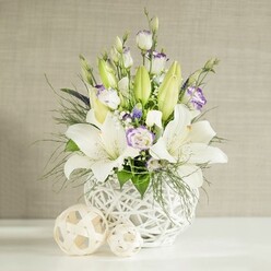A sophisticated wedding table arrangement is captured in gentle, diffused light, as if placed on a neatly dressed table in a North Watford reception venue. At its heart is a low, rounded white vase with an open lattice design that recalls woven wicker; the negative space in the pattern gives the whole piece a light, breathable quality. Rising gracefully from this base are prominent white lilies, some fully open with broad, velvety petals and clearly visible stamens, others still sheathed in elongated green buds that lend height and anticipation. Threaded between the lilies are stems of lisianthus, their buds and blossoms offering a softer, more romantic contrast: petals of creamy white subtly feathered with lavender-purple at the edges or clustered at the centre. Tiny white filler flowers, likely baby's breath, float among them like delicate clouds, while touches of deeper purple and blue blooms add small but impactful notes of colour. Foliage in multiple textures-fine, feathery sprigs, arching greenery, and broader leaves-fills out the design, gently cascading over the rim to give a natural, slightly untamed look, as if freshly gathered from a well-tended garden near St Albans Road. On the smooth white tabletop in front of the vase rest two decorative balls in pale beige tones, made from interlaced natural fibres that echo the vase's weave and complete the contemporary yet timeless aesthetic. The soft beige, horizontally textured background keeps the focus on the flowers while enhancing the sense of calm. Balanced between purity and quiet celebration, this piece would make a beautiful wedding centrepiece or a graceful gesture of comfort, easily delivered by local florists to homes and venues across North Watford.