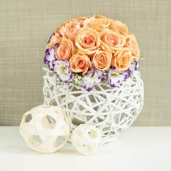 On a smooth white tabletop, a meticulously crafted centerpiece glows softly, as though ready to grace a wedding table in North Watford or a relaxed celebration near Garston. A lush, spherical mound of peach and apricot roses crowns the design, each bloom fully open and crowded closely together so that no stem is visible, only an expanse of velvety petals. Their colour moves gently from warm apricot in the depths to soft peach at the outer edges, lending the arrangement a subtle sunrise quality. Encircling the base of this rose dome is a ring of eustoma, their blossoms crisp and white but finely edged in a rich, royal purple that ripples along the ruffled petals. Sprinkled between them are tiny stars of white baby's breath, providing a delicate, cloud-like filler that keeps the arrangement from feeling heavy. All of this rests within a spherical white woven vase, created from interlaced wicker strands that twist and cross to form an open, airy lattice. Light passes through the gaps, throwing gentle patterns on the table. To one side sit two decorative wicker spheres in a pale natural wood shade, one medium and one smaller, their open weave mirroring the vase and tying the composition together. Behind, a soft beige fabric background with a subtle linen-like texture creates a calm, neutral setting. The overall feeling is tranquil and polished, ideal for couples in North Watford seeking a refined yet natural-looking centerpiece for a registry office reception, a restaurant gathering along St Albans Road, or a quiet anniversary dinner at home.