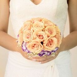 A detailed, chest-level image captures the midsection of a bride, her presence calm and composed, as she gently holds a round bridal bouquet in front of a pale, softly blurred background that could easily be the interior of a North Watford church or a light-filled community venue. Her classic wedding gown is a refined shade of white with an ivory undertone, the bodice featuring intricate lacework and a subtle V-neckline that flatters without distraction. The fabric around her waist is carefully draped, giving a wrapped, almost sculpted appearance that flows into the smooth, voluminous skirt. Resting securely in her hands-fingers gracefully curved, nails delicately manicured in a soft nude with a touch of shimmer on one thumb-is the focal point of the scene: a meticulously arranged bouquet of roses and accent blooms. The upper portion forms a compact dome of densely packed roses in shades of soft peach, light orange, and blush pink, their velvety petals layered in tight spirals that seem to glow gently, like early morning light over the rooftops near North Watford station. Beneath this pastel crown, a distinct border of deep purple flowers with frilled white edges, likely lisianthus, frames the base of the roses. This ruffled ring adds a rich, jewel-toned contrast, lending definition and sophistication to the design. Interspersed lightly around the lower edge, dainty white filler flowers similar to baby's breath introduce a fine, airy texture, softening the transitions between colours and giving the bouquet an almost cloud-like outline. The lighting is gentle and even, highlighting every curve of petal and lace. The image conveys romance, tradition, and thoughtful craftsmanship, perfectly suited to illustrate wedding bouquet ideas and colour palettes favoured by couples marrying in and around North Watford.