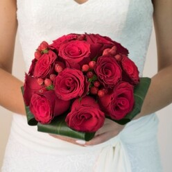 This evocative bridal bouquet, expertly styled by a North Watford florist familiar with local wedding traditions, is the very portrait of passionate elegance. The lens focuses closely on the bride-from her chest down-her immaculately manicured hands enveloping a sphere of densely packed, luxuriously red roses. Each rose is in varying stages of bloom, some petals curling gently outward while others form perfect spirals, a dynamic that suggests the unfolding story of a new marriage. Intermixed, vibrant hypericum berries pop with a radiant red gloss, their rounded shapes offering lively punctuation against the velvety calm of the roses. All are bounded at the base by broad, deep green foliage-shaped expertly into a collar that lends architecture and lushness, reminiscent of North Watford's scenic greenery. The bride's dress hints at classic sophistication, its white lace bodice and flowing lower fabric visible at the edge of the frame, juxtaposing the fiery bouquet. There's an unspoken aroma in the image: heady layers of rose mingling with the crispness of freshly cut leaves, as though plucked that morning from a North Watford market stall. Here, romance and tradition become one, in a bouquet that promises to be as memorable as a ceremony by Watford's storied avenues or community halls-an image filled with warmth, beauty, and the promise of lasting memories.