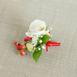 An elegant orchid boutonniere is photographed from above, lying on a light beige, woven fabric that resembles natural linen, its subtle texture creating a calm, neutral stage for the flowers. In the centre, a flawless white Phalaenopsis orchid opens with poised simplicity, its broad petals satin-smooth and gently curved, suggesting a sense of quiet devotion and care. At its core, touches of buttery cream and soft orange glow within the throat, offering warmth against the cool white. Surrounding the orchid, clusters of tiny white baby's breath blooms gather like little clouds, each minute star-shaped flower catching the soft light and giving the design a dreamy, romantic aura. Tucked just below and to the side is a vivid cluster of round, glossy red berries, reminiscent of hypericum, their polished skins adding a lively, almost jewel-like accent that feels particularly fitting for winter weddings or festive parties in and around North Watford's leafy streets. A few firm, pale green buds press in beside them, adding a hint of youthful freshness. Slender green leaves, smooth and gently tapering, weave through the arrangement, framing the orchid and berries with a touch of garden-like naturalness that calls to mind local parks such as Cassiobury. At the base, the stems are wrapped with a rich red satin ribbon, snugly wound and finished in a tidy knot whose short tails rest gracefully against the cloth. The composition feels intimate and considered, like a small token of love or celebration ready to be pinned to a lapel for a wedding, prom, or black-tie event in North Watford, carrying a quiet promise close to the heart.