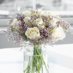 This image shows a thoughtfully composed sympathy bouquet, dominated by generous white and ivory roses that appear almost luminous against a veil of pale lavender filler flowers, like a pool of calm in the heart of North Watford. The roses are at perfect openness, their petals curling outwards in plush, layered spirals, each bloom distinct yet harmonising with its neighbours. Nestled gently around them are airy sprigs of tiny blossoms in soft lilac and blush-pink tones, creating a dreamy, cloud-like texture that softens the strong, rounded shapes of the roses. These filler flowers, likely statice or limonium, lend the arrangement an ethereal, almost weightless quality, as though it might float in the soft daylight streaming through a nearby window. The bouquet sits in a tall, slender clear glass vase filled with fresh water, the straight green stems clearly visible and neatly arranged, adding a sense of honesty and simplicity to the design. Resting on a white, uncluttered surface, the vase catches small reflections of light, while the background fades into a gentle blur of whites and light greys, suggesting a quiet interior somewhere off St Albans Road or near North Watford's leafy residential streets. Subtle daylight caresses the petals, highlighting their creamy undertones and creating delicate shadows that give the photograph depth without disturbing its calm. The mood is deeply peaceful and respectful, the kind of arrangement a local florist in North Watford might recommend for sending condolences, offering a tender, understated way to express sympathy, remembrance, and quiet support to a grieving family.