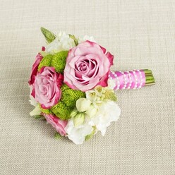 A compact, hand-tied bouquet rests gracefully on a neutral, light beige fabric that looks like fine linen, its subtle texture forming a calm backdrop that lets the flowers' colours sing. The arrangement is dominated by three lush pink roses, each bloom opening fully to reveal layered petals that curl inward at the center. Their colour moves gently from soft, powdery blush at the base of the petals to deeper pink and almost magenta at the tips, creating an elegant ombre effect that adds depth and romance. Between these roses, small round cushions of vivid lime green button chrysanthemums sit like playful accents, their countless tiny florets packed tightly together for a distinctly tactile, pom-pom appearance. Around and between the main blooms, cream and white filler flowers, resembling lisianthus or freesia, introduce a silky, feather-light texture that softens the whole composition. Delicate, starry white blossoms with fine yellow centers, reminiscent of wax flower, are scattered throughout, giving the bouquet a refined, almost lace-like finish. Wisps of dark and mid-green foliage peek from beneath the blossoms, offering contrast and a sense of natural movement without overwhelming the design. The stems are gathered into a short, tidy handle, wrapped in glossy fuchsia satin ribbon that echoes the roses' deeper tones. Over the ribbon, a narrow strand of white faux pearls is wound with care, crossing at angles like a tiny piece of jewellery for the bouquet. The trimmed stems extend slightly beyond the wrapping, showing fresh green ends. Bathed in soft, even light that emphasises freshness and gentle colour, the bouquet feels perfect for a North Watford bridesmaid walking from a ceremony near Garston to a reception in a local hotel, or as a delicate, modern-romantic centrepiece delivered to a dinner table by a trusted local florist.