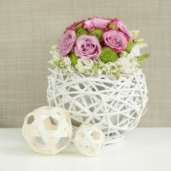 A round, white woven vase takes center stage on a spotless white surface, cradling a compact, dome-shaped bouquet that looks perfectly at home on a refined wedding table in North Watford. The flowers are tightly massed, with lush pink and lavender roses forming the main canopy; each blossom is open and generous, petals spiralling outward and showing hints of deeper rosy edges that create depth and warmth. Threaded between these roses are vivid green button chrysanthemums, their neat globes adding both texture and a refreshing, garden-bright contrast to the softer pastel tones. At the lower edge of the dome, small white and cream filler blooms tumble gently over, softening the transition between flowers and vase and adding a whisper of movement, as though a light breeze had just brushed past. The vase itself is made from thin, white twigs or rattan strands, woven into a spherical cage with visible gaps that let light through, giving the whole piece a modern, slightly Scandinavian feel while still echoing natural, handcrafted design often seen in local North Watford homes. To the left, two light-toned wicker balls, one large and one small, rest casually, echoing the basket weave and reinforcing the theme of organic materials. Behind everything, a softly blurred beige backdrop with a linen-like texture offers warmth without distraction. The image feels serene and composed, like a carefully styled table scene for a reception near St Albans Road, where fresh flowers and simple textures come together to create an atmosphere of relaxed elegance.