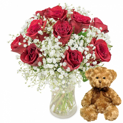 This image presents a classic romantic pairing styled with the quiet confidence of an experienced North Watford florist. On the left, a clear, gently curved glass vase cradles a generous bouquet of around a dozen deep red roses. Each rose is a rich, velvety crimson, the petals layered and plush, some blooms fully open, others still cupped, suggesting freshly cut stems ready for same-day delivery around North Watford. Interlaced among the roses are airy clusters of white baby's breath, their countless tiny blossoms forming a soft mist that lightens the intensity of the red and evokes the feeling of a tender whisper. The stems, visible through the vase, form straight green lines in the clear water, adding structure and a sense of honest simplicity. To the right of the flowers sits a medium-sized teddy bear in warm light brown plush, its fur appearing soft and slightly tousled, like a favourite childhood companion. The bear gazes forward with shiny dark eyes and a small stitched smile, while a dark brown ribbon is tied neatly at its neck, giving it a slightly dressed-up charm. Everything is set against a clean, bright white background that eliminates distraction and allows the colours and textures to stand out: the satin sheen of the petals, the airy froth of gypsophila, the smooth transparency of glass, and the cosy pile of the bear's fur. This carefully balanced scene suggests a thoughtful gift for a partner in North Watford, perfect for birthdays, anniversaries, or a spontaneous romantic gesture that feels both traditional and deeply personal.