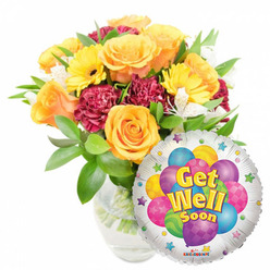 This cheerful "Get Well Soon" arrangement captures a hospital-bed uplift and a neighbourhood visit all at once: a full bouquet in a clear glass vase fills the left side of the scene, while a round silver Mylar balloon rests to the lower right. The bouquet is built around several large orange and warm yellow roses, their soft velvety petals unfurling into open, reassuring blooms; you can almost sense a faint citrusy-sweet scent in the description. Bright yellow gerbera daisies provide a lively counterpoint with their flat golden faces, and deep burgundy or maroon carnations give weight and ruffled texture, creating a rich visual rhythm. Sprigs of white alstroemeria peek through as delicate, airy highlights, and a variety of glossy green foliage cradles the stems; the clear vase reveals water and neatly trimmed stems, evidence of careful florist technique. The metallic balloon's surface reflects daylight and showcases a playful central graphic of cartoon balloons in purple, blue, green, pink and yellow-some patterned with stripes or dots-while bold yellow-and-blue letters say "Get Well Soon" with a subtle shadow for emphasis; a tiny "KALEIDOSCOPE" logo is visible along the bottom edge. As someone who arranges flowers for neighbours between Watford High Street and North Watford station, I imagine this pair delivered with a handwritten note: it reads as encouragement, friendly care, and a little local sunshine, an ideal offering for recovery, hospital visiting, or a bedside brightener from Flower Delivery North Watford.