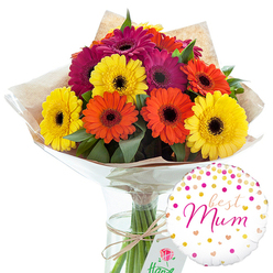 In this carefully composed image, a cheerful bouquet of gerbera daisies is presented as if ready to be carried out of a North Watford florist and straight into a loved one's home. The arrangement is dominated by generously sized gerbera heads in a harmonious blend of sunny yellow, bright tangerine orange, and sumptuous deep magenta, each flower displaying its signature ring of flat petals around a contrasting darker centre. The blooms sit closely together, forming a rounded, almost dome-like shape, while pockets of lush, dark green foliage peek out between them, adding texture and a feeling of natural abundance. The stems are gathered by hand and wrapped in a layer of softly crumpled brown kraft paper, giving a rustic, market-fresh look reminiscent of flowers picked up near Cassiobury Park or the local shops off St Albans Road. Over this sits a clear, glossy cellophane wrap that reflects the studio lighting with gentle highlights, hinting at the bouquet's crisp freshness. A length of plain twine is tied around the stems in a neat bow, balancing simplicity with care. The bouquet stands upright in a slender, transparent glass vase filled with clean water, making the green stems a key part of the visual composition. To the lower right, a round helium balloon in bright white adds a festive note; it is speckled with confetti-like dots and tiny hearts in shades of gold and pink, with the affectionate words best Mum scripted in pink at its centre. Together, flowers and balloon create an image of warmth, gratitude, and celebration, perfect for honouring mothers and mother figures across North Watford.