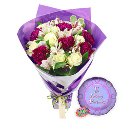 Set against a clean white backdrop, this elegant romantic gift from a North Watford florist pairs a lush hand-tied bouquet with a coordinating balloon. The flowers form a rounded, full arrangement dominated by creamy white roses, each bloom classically shaped with layered, unfurling petals that hint at a gentle fragrance. Woven between them, rich magenta and burgundy carnations bring depth and drama, their ruffled heads packed with silky petals that create a soft, cloud-like texture. White alstroemeria, or Peruvian lilies, add a lighter, more intricate note: their slender petals, etched with fine reddish-brown stripes and speckles, feel almost like watercolour brushstrokes. Around the blooms, glossy green leaves and touches of variegated foliage frame the composition, giving it a fresh-cut-from-the-garden impression, reminiscent of walking home from the shops along St Albans Road with flowers in hand. The bouquet is wrapped with care in a double layer: crisp white paper close to the stems for structure, and sheer, crinkled purple cellophane forming a luminous collar that rises behind the flowers and reflects soft violet highlights. At the gathered stems, a bright lime-green satin ribbon is tied into a neat bow, holding a small tan gift tag that seems ready for a personal love note. To the right of the bouquet lies a round metallic purple foil balloon with a scalloped edge, echoing the shape of a prize rosette. At its centre, set against a paler lavender circle, the phrase "To Loving Partners" is written in flowing, romantic script, accompanied by a tiny rainbow holographic label that catches the light. This coordinated set feels ideal for anniversaries, proposals, or Valentine's surprises, delivered straight to doors across North Watford.