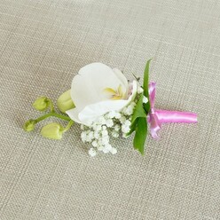 A single white Phalaenopsis orchid, immaculate and softly luminous, forms the centrepiece of this handcrafted boutonniere, arranged as if ready to pin onto a suit lapel for a North Watford wedding or evening event. It rests horizontally on a light beige linen-textured fabric, whose fine, woven surface adds a quiet, natural backdrop, reminiscent of a freshly pressed jacket laid out before a special day. The orchid's broad petals open in a smooth, elegant curve, pure white at the edges and warming to a gentle wash of creamy yellow and the faintest pink tones at the throat, where the intricate inner structure is just visible. Around the base of the bloom, a halo of white baby's breath gathers in small, cloud-like clusters, their tiny rounded flowers creating a soft, romantic haze that lightens the whole design. A few slim, fresh green leaves frame the composition from behind, their colour bringing to mind the leafy streets and gardens scattered through North Watford. Extending from one side, a short stem holds several lime-green orchid buds, still closed and glossy, hinting at new beginnings and the unfolding of the day ahead. The stems are neatly wrapped in a vivid fuchsia pink satin ribbon, tied into a compact bow with short, tidy tails; the ribbon's bright, jewel-like colour feels joyful and modern, ideal for couples wanting a small pop of personality beside a classic suit. Soft, even lighting glides across the petals, the baby's breath, and the linen, highlighting their different textures and giving the entire floral accessory a sense of poised calm and heartfelt intention, suitable for grooms, best men, or special guests across local venues and celebrations.