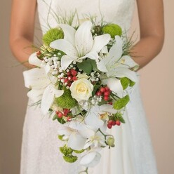 As sunlight filters softly through the paneled windows of a North Watford venue, a bride cradles an exquisite, cascading bouquet-a masterwork of local floral artistry. Dominating this arrangement are luminous white lilies, their elongated petals tinged with the barest hint of bronze speckles that catch the light and draw you deeper into their silken centers. Nestled among them is a luscious cream rose, its spiral of petals tight and hopeful. The bouquet overflows with texture: plump lime-green pom flowers bring a burst of energy, while trailing tendrils of white Phalaenopsis orchids sweep downward, bestowing a sense of gentle movement. Fluffy clouds of tiny baby's breath lighten the whole and offer subtle contrast to the rich red berries-clusters of Hypericum scattered artfully to symbolize joy and prosperity. Delicate sprays of greenery and the occasional arching blade frame the flowers, enhancing the natural feel treasured by North Watford's discerning brides. Against the blurry lace of a wedding gown, the composition feels both timeless and on-trend, capturing the anticipation and beauty of a memorable local ceremony. Each bloom appears hand-chosen, each wound ribbon and leafy accent arranged with the skilled touch of a North Watford florist devoted to making wedding dreams blossom.