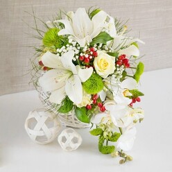 An opulent cascading arrangement, photographed on a pristine white tabletop against a muted beige, fabric-like background, captures the quiet elegance of a modern wedding reception in North Watford. The flowers are gathered into a low, round white wicker basket whose textured weave adds a natural, handcrafted touch. Dominating the design are several large white lilies, their trumpet blooms fully open, with curved petals, visible stamens, and subtle speckling that lends depth and refinement. At the upper centre, a single cream-yellow rose is poised between bud and bloom, its velvety petals spiralling inward like a soft focal jewel. From the right side of the main cluster, stems of white Phalaenopsis orchids trail downward in a gentle arc, creating a graceful cascade that suggests a waterfall bouquet often chosen by brides along St Albans Road and nearby venues. Lush greenery surrounds and supports the blooms: fine, wispy grasses, fern-like fronds, and broader glossy leaves that frame the lilies and rose. Bright green, rounded pom-pom flowers reminiscent of green trick dianthus nestle between the leaves, bringing a playful, tactile texture. Tiny sprays of white baby's breath float among the petals, while clusters of glossy red berries punctuate the arrangement with bursts of rich colour, evoking a festive, seasonal mood. To the left of the basket, two decorative white woven spheres, one larger and one smaller, echo the wicker texture and add a sculptural, minimalist accent. The entire composition, crafted with the care you'd expect from an experienced North Watford florist, radiates serenity, freshness, and an understated luxury perfect for a top table or intimate dinner setting.