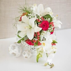 On a clean white tabletop, an elegant floral centrepiece is presented with the quiet confidence of a professionally styled wedding table, the sort you might find in a boutique venue tucked off St Albans Road in North Watford. The arrangement sits in a round white basket-style container, its open, criss-crossed weave creating a lace-like cage around the stems while letting the light play through the gaps. From this airy base rise dramatic white Oriental lilies, their broad, satin-like petals flaring open to reveal golden pollen-dusted stamens, suggesting a soft, heady perfume. Threaded among them are rich red roses, their velvety petals spiralling inward in snug, romantic cups, each one a deep note in the bouquet's visual melody. Wisps of white baby's breath drift between the dominant blooms like tiny clouds, while fine, bright green fern-like foliage and other fresh greenery add movement and keep the composition feeling light rather than heavy. Small clusters of shiny red hypericum berries are dotted throughout, introducing playful pops of colour that echo the roses and hint at festive gatherings or winter weddings. One side of the design is graced by a cascading stem of white Phalaenopsis orchids that spill gently over the edge, each orchid bloom displaying a subtle yellow and pink throat, with a few buds still waiting to open. To the left of the main basket, three white woven spheres in varying sizes rest on the table, mirroring the basket's texture and giving the scene an artful, curated feel. The neutral beige background remains soft and unobtrusive, ensuring all attention is drawn to the interplay of white, red, and green. The arrangement feels both luxurious and welcoming, ideal as a North Watford wedding table arrangement, a Christmas-season centrepiece, or a refined gift delivered for an intimate celebration.