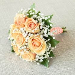 Capturing the sophisticated charm of a local North Watford wedding, this visually arresting bouquet is composed of seven or eight sumptuous, fully bloomed roses. Their hues drift gracefully between blush peach and sunlit apricot, each unfurling petal illuminated by the soft northern daylight typical of Watford's peaceful side streets. Artfully dispersed among these roses, soft tufts of white baby's breath hover, granting the arrangement a floating, cloudlike quality that feels both delicate and uplifting. Deep green, glossy rose leaves fan discreetly out between the florals, grounding the bouquet and adding that unmistakable touch of authentic garden greenery found so often in North Watford's hidden corners. The stems are held together with a coral satin ribbon, embellished in a gentle spiral by dainty white pearls-an echo of classic elegance and bridal refinement. Resting atop a neutral linen surface, the bouquet's rounded, tailored shape and shimmering accents invite thoughts of love-filled ceremonies at local venues or garden receptions. One can almost sense a trace of fresh floral fragrance in the air, evoking emotions of joyful anticipation and quiet sophistication-a bouquet designed to stand out at life's most cherished milestones.