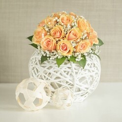 An elegant floral centerpiece glows against a softly textured, light beige wall, the kind of arrangement you might find gracing a carefully styled home in North Watford or a boutique hotel lobby off St Albans Road. At the core is a generous dome of approximately twenty to thirty peach roses, their petals tightly layered yet just beginning to unfurl. Each bloom transitions from a deeper, amber-orange heart to lighter, almost blush edges, creating a gentle ombré effect that feels warm and inviting. Interwoven throughout the roses are fine sprays of white baby's breath, their tiny blossoms forming a mist-like halo that softens the overall profile. Here and there, dark green leaves emerge beneath the flowers, adding depth, natural contrast, and a hint of woodland freshness, as though the bouquet has just been gathered from a sheltered garden. The entire floral crown rests in a large, round white vase constructed from intricately woven strips that form an open lattice. This sculptural vessel has the appearance of delicate wicker or rattan yet feels modern and minimal, its crisp white tone framing the flowers without competing for attention. On the smooth white tabletop in front of the vase sit two smaller woven spheres in light beige or cream, their airy, open structures echoing the main container's design and adding a sense of playful balance to the composition. Soft, even lighting washes over the arrangement, picking up the satin sheen of the rose petals and the gentle shadows cast by the woven elements. The scene conveys quiet sophistication and would be perfectly at home as a wedding centerpiece, a showpiece for home staging, or a refined gift arrangement curated by a local North Watford florist for same-day delivery across the area.