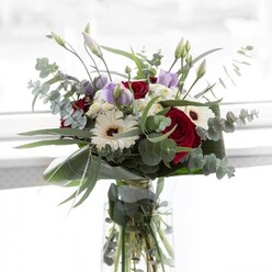 Bathed in the gently filtered daylight common to North Watford's sunlit terraces, this bouquet possesses a refined sophistication that captures both tradition and freshness. Rich, velvety red roses dominate the arrangement, their deep hues softened by the pure white gerbera daisies, each bloom unfurling with a texture reminiscent of handcrafted paper. The bouquet is punctuated by sprigs of eucalyptus, their round, silvery leaves arching elegantly, offering both structure and a cool, invigorating fragrance familiar to those who stroll along Chalk Hill. Interlaced among the bolder blooms, delicate mauve lisianthus buds-some just beginning to open-introduce an air of anticipation and gentle promise, perfect for moments of remembrance or celebration unique to the North Watford community. Fluffy white filler flowers add a nuanced cloud-like volume, echoing the gentle rhythm of quiet Saturday mornings at local markets. The vase's transparency draws the eye to robust, verdant stems, a testament to the bouquet's radiant freshness. Every stem seems thoughtfully nestled, each flower in companionable harmony, creating an arrangement equally suited for brightening a kitchen table on Queens Road or gifting as a heartfelt gesture on important local occasions.