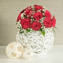 At the heart of this scene is a rounded bouquet of sumptuous red roses, their petals dense and velvety, spiralling inward with the kind of fullness that suggests they have just been arranged by a careful florist's hand. Each rose is at its peak, cupped closely against its neighbours so that the blooms form a smooth, dome-like silhouette. Threaded delicately between them are airy sprays of white baby's breath, the tiny star-shaped flowers creating a gentle halo around the roses and softening their intensity with a whisper of lightness. Here and there, discreet touches of green foliage emerge, grounding the composition in something fresh and alive, as if it were set on a wedding table at a North Watford hotel near Garston or along St Albans Road. The flowers rest inside a striking white vase, bulbous and rounded, built from slender strands that loop around each other into a loose, open-weave structure. This design lets light and shadow slip through, giving the vase an almost sculptural presence that complements the roses' opulence. In front of it, on a smooth, pale surface, sit two creamy, woven spheres, one larger and one smaller, both echoing the same latticework and adding a modern decorative touch. The background is a soft blur of warm neutral tones, like a textured wall lightly brushed with beige, keeping the focus firmly on the depth of the reds and the crisp whites. The overall impression is one of quiet luxury and romance, a centrepiece worthy of a wedding breakfast or anniversary dinner in North Watford, arranged with the kind of artistry that makes guests pause and admire.